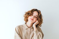 Young Caucasian woman with wavy bob hairstyle and natural makeup wearing beige oversized shirt poses dreamily against white background, hand on cheek.
