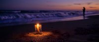 Glowing lantern on sandy beach at sunset with silhouette of person walking by ocean waves. Romantic evening atmosphere for travel and relaxation concepts.