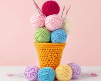 Colorful yarn balls arranged in crocheted yellow cone basket with knitting needles on pink background, creative crafting composition for knitting and crochet projects.