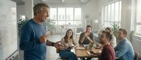 Middle-aged male with gray hair and light skin tone, wearing casual blue shirt, leading business meeting or training session with diverse group of colleagues in modern office environment with natural lighting.