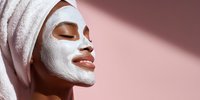 Young African woman enjoying spa treatment with white clay facial mask and towel on head against pink background for skincare and wellness routine.