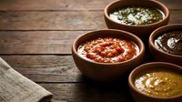 Four rustic clay bowls filled with colorful sauces on a dark wooden table, showcasing traditional condiments for cooking and dining.