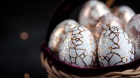Decorative white eggs with elegant crackle pattern in golden metallic finish nestled in dark basket against black background with soft bokeh lights.