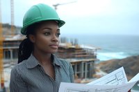 Young African American female construction engineer in green hardhat reviewing blueprints at coastal construction site, wearing striped business shirt against ocean background.