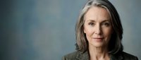 Middle-aged woman with long gray hair and fair complexion wearing a gray blazer, smiling warmly at camera against soft blue background for corporate or professional use.