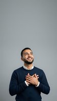 Middle-aged man with dark hair and beard, South Asian ethnicity, wearing navy sweater over collared shirt, hands on chest, looking upward with grateful expression against neutral gray background.