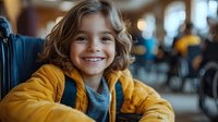 Young child with wavy brown hair wearing yellow jacket and gray shirt smiles brightly at camera, showing genuine happiness and joy in casual indoor setting with blurred background.