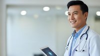 Professional Asian male doctor smiling while holding digital tablet in modern hospital setting with stethoscope around neck for healthcare technology concepts.