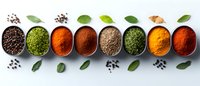 Colorful aromatic spices and herbs in metal bowls arranged in row on light background with scattered peppercorns and bay leaves for cooking and seasoning.