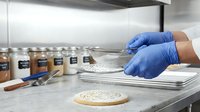 Scientist in blue latex gloves examining flour sample with metal spoon in laboratory kitchen setting with ingredient jars and baked goods for food testing and quality control research.