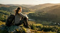 Woman hiker with backpack sitting on mountain cliff overlooking forested valley and river during golden hour, outdoor adventure and nature exploration.