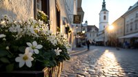 Charming European cobblestone street at sunset with white flowers in window box, church tower in background, golden sunlight reflecting on ancient pavement.