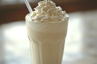 Creamy vanilla milkshake topped with swirls of whipped cream served in tall glass with straw, soft focus background creates dreamy atmosphere for food photography.