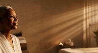 Middle-aged Black woman in white bathrobe relaxing in spa bathroom with warm golden lighting and window shadows, wellness and self-care concept.