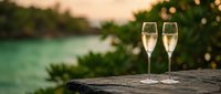 Two champagne flutes filled with sparkling wine on stone ledge overlooking turquoise tropical lagoon at sunset with palm trees and warm golden light.