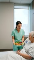 Young Hispanic female nurse in green scrubs serving healthy meal to elderly Caucasian patient in modern hospital room for nutrition care and recovery.