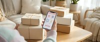 Young woman with light skin tone, wearing casual clothing, holding smartphone displaying delivery tracking app surrounded by cardboard boxes on wooden table, representing e-commerce logistics and online shopping management.