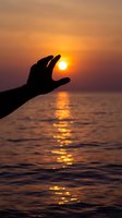 Hand silhouette reaching toward golden sunset over ocean water with warm orange sky and sun reflection creating peaceful spiritual moment.