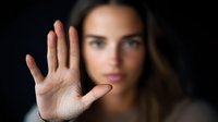 Young woman with serious expression showing stop gesture with palm against dark background, focus on raised hand in foreground.