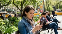 Young woman using smartphone while sitting on park bench in urban setting with trees and city street background.