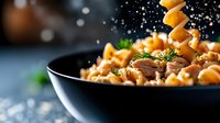 Delicious pasta with mushrooms and herbs in black pan with falling cheese cubes and seasoning creating dynamic cooking scene for culinary content.