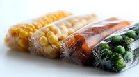 Fresh frozen vegetables in transparent vacuum sealed bags, including corn, carrots and broccoli florets arranged in rows on white surface with soft lighting.