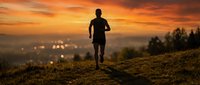 Male athlete jogging on rural hillside at sunset with city lights glowing below. Silhouetted runner training outdoors during golden hour for fitness, wellness, and active lifestyle concepts.