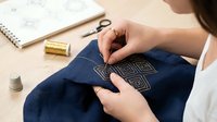 Woman hands embroidering geometric pattern on dark blue fabric with golden thread, traditional needlework craft hobby at home workspace with sewing supplies.