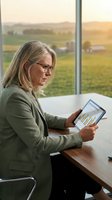 Mature Caucasian businesswoman analyzing financial data on tablet in modern office with countryside view during golden hour sunset.