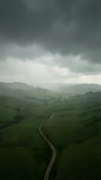 Winding road through lush green rolling hills under dramatic stormy sky, aerial view of rural landscape with mountains in distance, suitable for travel and nature backgrounds.