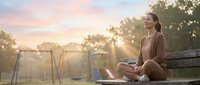 Young Asian woman practicing mindfulness meditation on park bench during golden hour sunset with peaceful expression and crossed legs pose.