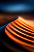 Abstract macro photography of curved orange metallic surface with dramatic lighting and deep blue background creating dynamic contrast and flowing lines.