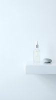 Clear facial serum or essence in glass dropper bottle with silver cap displayed on white platform beside smooth stone for skincare and cosmetic product marketing.