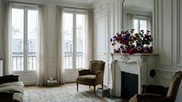 Elegant Parisian apartment living room with ornate fireplace, vintage armchairs, tall windows, and vibrant floral arrangement in classic white interior.