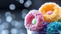 Colorful frosted donuts with sprinkled sugar coating on dark background with bokeh lights. Sweet dessert food photography with vibrant pink, yellow and blue colors.