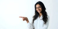 Young South Asian woman in white lab coat pointing to side with confident smile against clean white background for healthcare or professional concept.