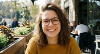 Young woman with glasses and curly brown hair smiling at outdoor cafe terrace. Casual portrait of happy female in yellow sweater enjoying sunny day at restaurant.