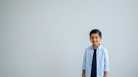 Young Asian male child wearing light blue button-up shirt and dark tie, smiling at camera against neutral white background, suitable for educational materials, business concepts, and professional stock use.