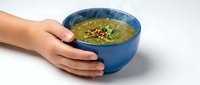 Hands holding blue ceramic bowl with green soup garnished with herbs and spices on white background for healthy eating and cooking concepts.