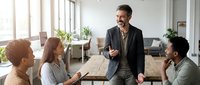 Middle-aged male professional with dark hair and beard, Mediterranean ethnicity, wearing business casual attire, leading a team meeting with three diverse colleagues in a modern office environment with natural lighting and plants.