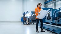 Engineer in orange safety vest and glasses reviewing blueprints at industrial manufacturing facility with machinery and workers in background for factory operations and construction planning.