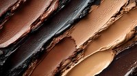 Diagonal smears of creamy foundation makeup in different skin tone shades from light beige to deep brown showing texture and cosmetic swatches on dark background.