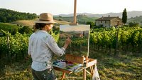 Artist painting vineyard landscape on canvas at easel during golden hour in Tuscany countryside with rolling hills and stone buildings.