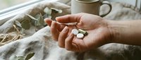 Woman's hand holding white chamomile flowers on linen fabric with dried herbs and ceramic cup, natural wellness and herbal tea preparation.