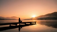 Person meditating on wooden dock at sunrise over misty mountain lake with golden light reflection creating peaceful zen atmosphere for wellness content.