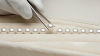 Gloved hand holding a pearl with tweezers over a lustrous necklace on cream velvet, showcasing jewelry craftsmanship and quality control.