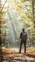 Hiker with backpack standing on autumn forest path surrounded by falling leaves and golden foliage, exploring nature during fall season.