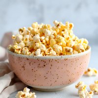 Fresh buttery popcorn served in pink ceramic speckled bowl on light marble background with scattered kernels and soft natural lighting.