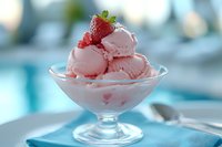 Pink strawberry ice cream scoops in glass dessert bowl topped with fresh berry, served on blue table setting against soft blurred background.