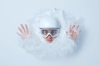 Young Caucasian man in ski goggles and protective helmet emerging through deep powder snow, raising hands in playful gesture against white winter background.